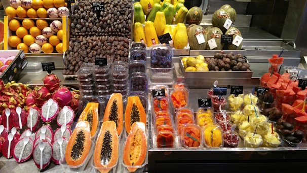 A vibrant display of fruits at a market, featuring an array of colorful produce including dragon fruits with spoons, papayas, melons, dates, and assorted fruit slices neatly packaged. Price tags are visible, indicating a commercial setting with various types of fresh and possibly exotic fruits.