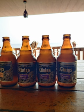 Four amber-colored beer bottles are placed on a wooden table. The bottles have a detailed label with the word 'Kings' and are arranged in a row. In the background, there's a blurred view of an outdoor setting with a railing and a body of water, likely the ocean or a sea. The ceiling is visible with a pendant light hanging down.