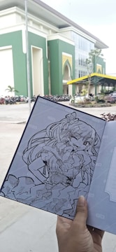 A hand is holding a notebook with a detailed black and white drawing of an anime-style character. The background shows a large, modern building with green and white walls, an arched entrance, and a parking area with numerous parked motorcycles.