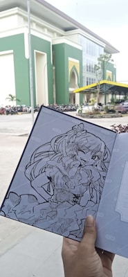 A hand is holding a notebook with a detailed black and white drawing of an anime-style character. The background shows a large, modern building with green and white walls, an arched entrance, and a parking area with numerous parked motorcycles.