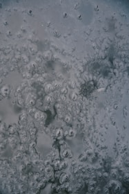 Patterns of bubbles trapped in ice are visible, featuring intricate, swirling designs. The surface appears cold and smooth, with varying shades of gray and subtle hints of green.