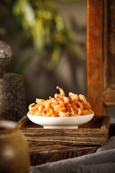 A white ceramic bowl filled with dried shrimp placed on a wooden tray. The tray rests on a fabric surface, and the background includes blurred natural elements and stone textures, suggesting a rustic or natural setting.