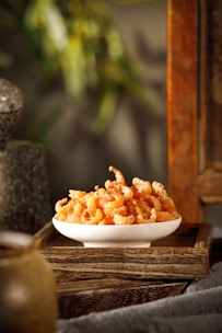 A white ceramic bowl filled with dried shrimp placed on a wooden tray. The tray rests on a fabric surface, and the background includes blurred natural elements and stone textures, suggesting a rustic or natural setting.