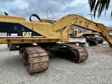 Several large yellow excavators with 'CAT' branding are parked on a gravel surface under a cloudy sky. The machines feature large metallic tracks and articulated arms, typically used for heavy-duty digging and construction work.