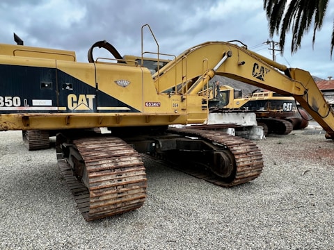 Several large yellow excavators with 'CAT' branding are parked on a gravel surface under a cloudy sky. The machines feature large metallic tracks and articulated arms, typically used for heavy-duty digging and construction work.