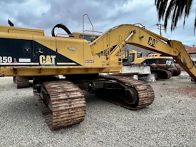 Several large yellow excavators with 'CAT' branding are parked on a gravel surface under a cloudy sky. The machines feature large metallic tracks and articulated arms, typically used for heavy-duty digging and construction work.