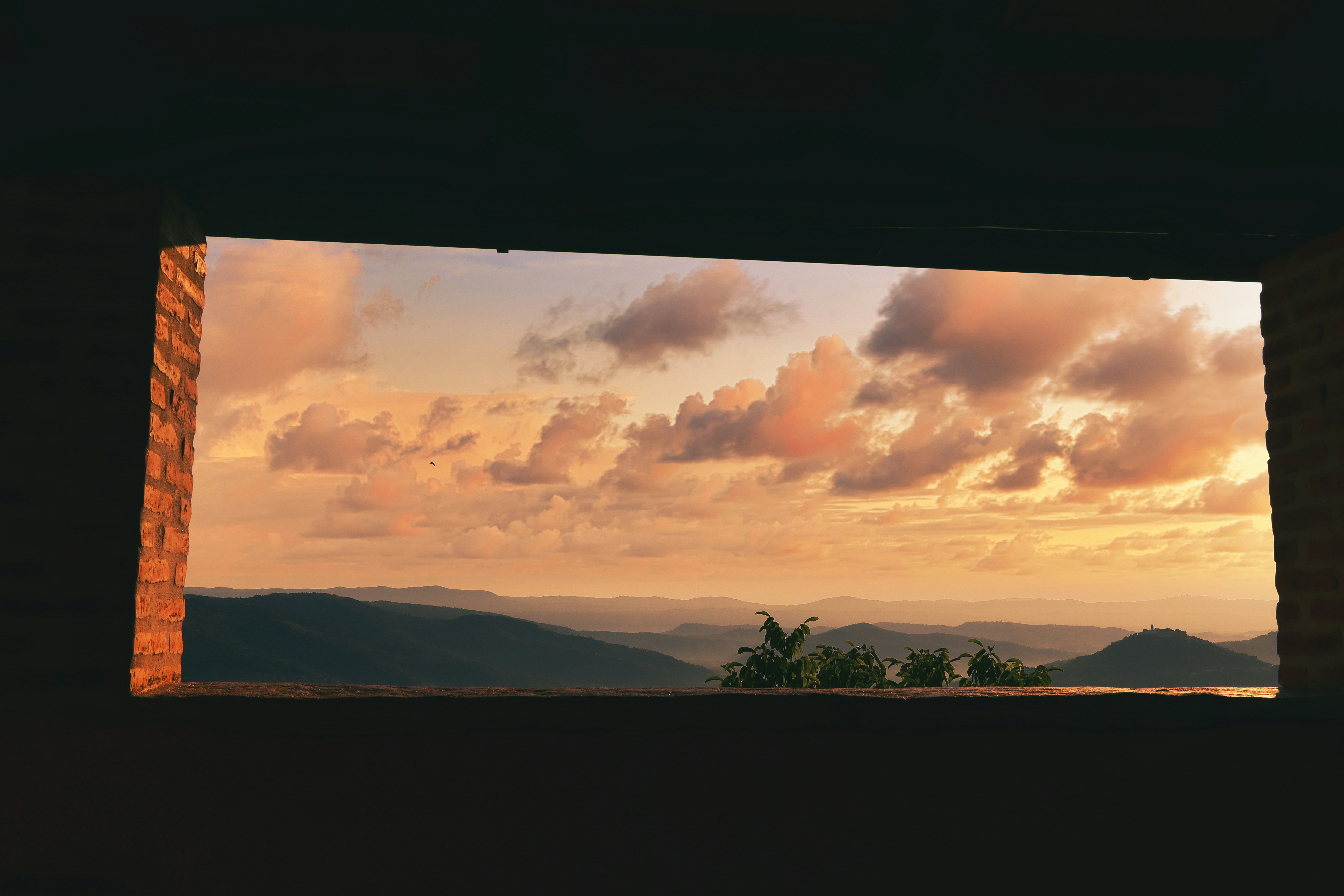 Interior window frames a sunset landscape with orange clouds over distant mountains, while a small cluster of palm-like trees sits on the horizon.