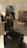A seated black stone statue depicting an Egyptian pharaoh with a traditional headdress, displayed in a museum setting. In the background, there is a colorful checkerboard patterned artifact enclosed in glass, and several people are observing other exhibits.