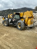 A construction site featuring a large yellow and black telescopic handler with large wheels. The equipment is positioned on a dirt path with tire tracks visible in the foreground. In the background, there are rocky hills and a cloudy sky, adding an overcast atmosphere to the setting.
