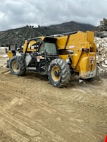 A construction site featuring a large yellow and black telescopic handler with large wheels. The equipment is positioned on a dirt path with tire tracks visible in the foreground. In the background, there are rocky hills and a cloudy sky, adding an overcast atmosphere to the setting.