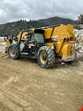 A construction site featuring a large yellow and black telescopic handler with large wheels. The equipment is positioned on a dirt path with tire tracks visible in the foreground. In the background, there are rocky hills and a cloudy sky, adding an overcast atmosphere to the setting.