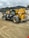 A construction site featuring a large yellow and black telescopic handler with large wheels. The equipment is positioned on a dirt path with tire tracks visible in the foreground. In the background, there are rocky hills and a cloudy sky, adding an overcast atmosphere to the setting.