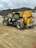 A construction site featuring a large yellow and black telescopic handler with large wheels. The equipment is positioned on a dirt path with tire tracks visible in the foreground. In the background, there are rocky hills and a cloudy sky, adding an overcast atmosphere to the setting.