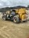 A construction site featuring a large yellow and black telescopic handler with large wheels. The equipment is positioned on a dirt path with tire tracks visible in the foreground. In the background, there are rocky hills and a cloudy sky, adding an overcast atmosphere to the setting.
