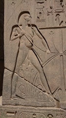 A detailed ancient Egyptian stone carving depicting a figure with a tall headdress holding a spear. Hieroglyphs surround the figure, and there are intricate details across the entire stone surface.