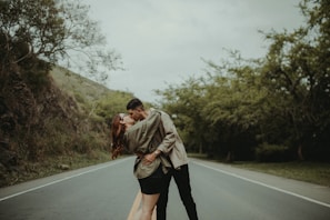 A couple embraces in a romantic kiss on an empty road surrounded by greenery, with lush trees on either side. The atmosphere is serene, enhanced by the overcast sky above.