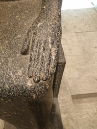 A close-up view of a sculpture's hand resting on a textured stone surface. The hand is intricately carved with visible speckles and a rough texture typical of ancient stone artwork.