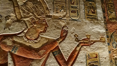 A detailed carved relief depicts an ancient Egyptian figure adorned with a headdress, holding a ceremonial object. The figure is intricately etched into a stone wall, surrounded by hieroglyphics and symbols.
