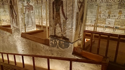 Located within an ancient tomb, the scene features hieroglyphic inscriptions and detailed wall paintings depicting Egyptian deities. The area is constructed with stone and preserved with added support beams, illuminated with soft, warm lighting that highlights the intricate artwork.