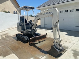 A small excavator is parked on a concrete driveway in front of a garage. The machine is primarily white with some black components and appears to be in a residential area. The background includes two houses with beige walls and tiled roofs. It is a sunny day, as evidenced by the strong shadows and the bright lighting.