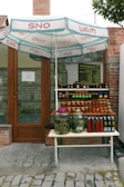 A small outdoor stall is set up in front of a brick building with a wooden door. Various jars of preserved vegetables and sauces are neatly displayed on a two-tiered table. Two large jars contain pickled items, and there are several smaller jars and bottles with labels. A large umbrella with blue and red lettering provides shade over the setup.