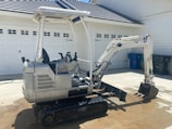 A small excavator is parked on a concrete driveway in front of a garage with white doors. The machine has a silver and white body with a black seat and digger arm, and is equipped with rubber tracks. The background features recycling bins.