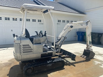 A small excavator is parked on a concrete driveway in front of a garage with white doors. The machine has a silver and white body with a black seat and digger arm, and is equipped with rubber tracks. The background features recycling bins.