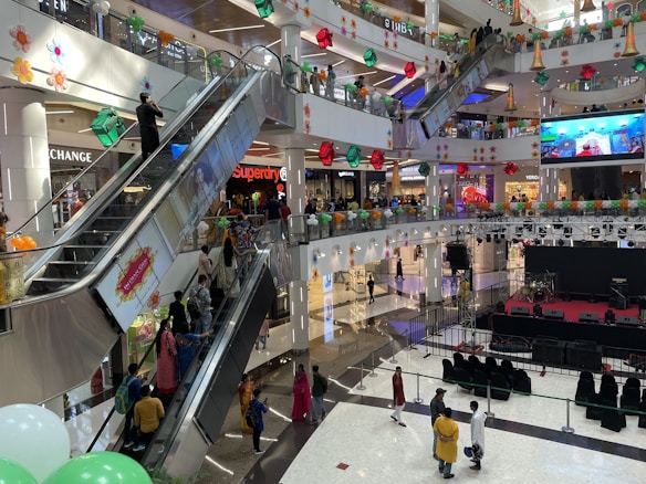 A bustling shopping mall with multiple floors filled with people moving on escalators and walking around. The area is adorned with colorful decorations, including flowers and geometrical shapes. Stores like Superdry and other fashion outlets are visible, and there is a stage setup with musical equipment.