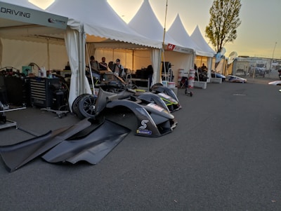Several white tents are set up on a paved surface at what appears to be a race track pit area. Under the tents, various car parts, tools, and equipment are visible. There are a few people working on or standing near racing cars. The setting sun casts a warm glow, creating long shadows. A tree is visible in the background along with fencing and additional parked vehicles.