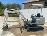 A small excavator with a white and grey color scheme is parked on a concrete surface. The excavator has a rubber track undercarriage and an open cab with a seat and control levers. Behind it, a light-colored wall and a tile-roofed building are visible, along with some green foliage including palm trees.