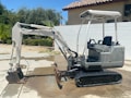 A small excavator with a white and grey color scheme is parked on a concrete surface. The excavator has a rubber track undercarriage and an open cab with a seat and control levers. Behind it, a light-colored wall and a tile-roofed building are visible, along with some green foliage including palm trees.