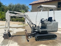 A small excavator with a white and grey color scheme is parked on a concrete surface. The excavator has a rubber track undercarriage and an open cab with a seat and control levers. Behind it, a light-colored wall and a tile-roofed building are visible, along with some green foliage including palm trees.