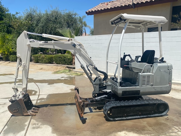 A small excavator with a white and grey color scheme is parked on a concrete surface. The excavator has a rubber track undercarriage and an open cab with a seat and control levers. Behind it, a light-colored wall and a tile-roofed building are visible, along with some green foliage including palm trees.