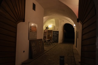 A dimly lit, narrow alleyway with cobblestone pavement and curved archways. On the left, a sign indicating a wine shop with small tables and chairs in front of it. The warm yellow glow of lights creates an inviting ambiance.