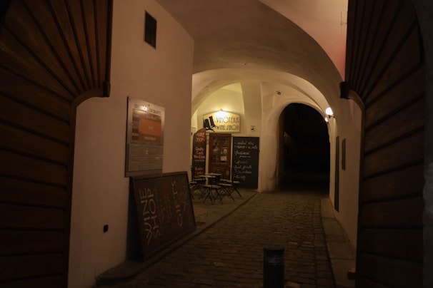 A dimly lit, narrow alleyway with cobblestone pavement and curved archways. On the left, a sign indicating a wine shop with small tables and chairs in front of it. The warm yellow glow of lights creates an inviting ambiance.