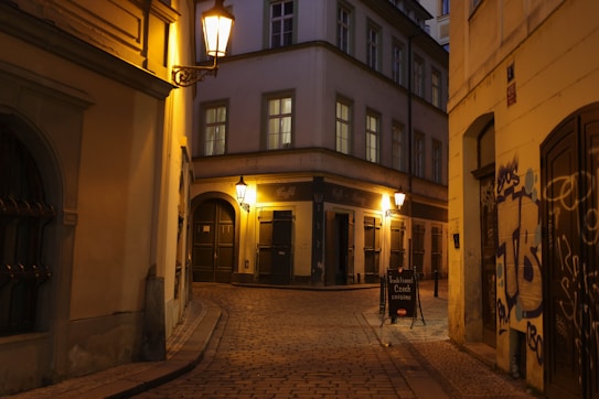 A dimly lit, narrow cobblestone street curves between historical buildings. Warm yellow street lamps cast a soft glow against the architecture, highlighting details like windows and doorways. A sign advertising 'Traditional Czech Cuisine' stands on the sidewalk. Some graffiti decorates a wall, and the scene suggests quietness with no visible people.