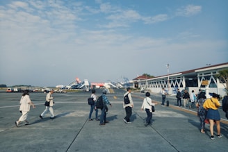 Several people walk across the tarmac at an airport, heading towards a terminal. Multiple aircraft boarding stairs are visible in the background, alongside a building that appears to be part of the airport infrastructure. The sky is partially cloudy, and the scene conveys a sense of movement and travel.