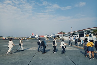 Several people walk across the tarmac at an airport, heading towards a terminal. Multiple aircraft boarding stairs are visible in the background, alongside a building that appears to be part of the airport infrastructure. The sky is partially cloudy, and the scene conveys a sense of movement and travel.