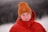 A person wearing a warm orange knit beanie and a red puffer jacket stands outdoors in a snowy environment. The background is blurred, emphasizing the focus on the individual.