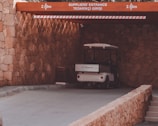 A small white utility vehicle is parked under a stone archway marked with height restrictions, leading to a suppliers' entrance. The surroundings are constructed from stone, with neutral tones and a rustic appearance.