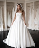 A smiling woman wearing a strapless white wedding dress and veil stands in a bridal boutique surrounded by other dresses hanging on display.