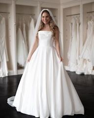 A smiling woman wearing a strapless white wedding dress and veil stands in a bridal boutique surrounded by other dresses hanging on display.
