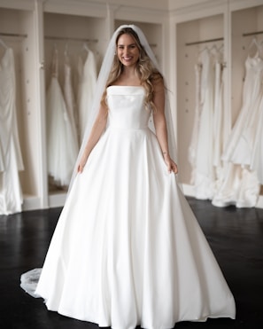 A smiling woman wearing a strapless white wedding dress and veil stands in a bridal boutique surrounded by other dresses hanging on display.