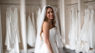 A smiling bride in a white strapless wedding dress with a veil stands in front of a row of hanging wedding gowns in a boutique or dressing room.