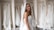 A smiling bride in a white strapless wedding dress with a veil stands in front of a row of hanging wedding gowns in a boutique or dressing room.