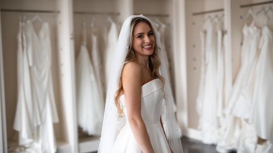 A smiling bride in a white strapless wedding dress with a veil stands in front of a row of hanging wedding gowns in a boutique or dressing room.
