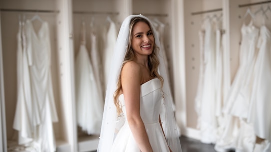 A smiling bride in a white strapless wedding dress with a veil stands in front of a row of hanging wedding gowns in a boutique or dressing room.