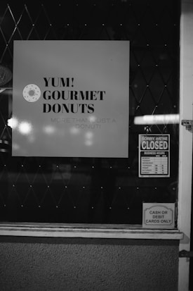 A window display featuring a sign with 'YUM! Gourmet Donuts' and the phrase 'More than just a donut!' is visible. Nearby, there is a sign indicating the store is closed with business hours listed. Another sign states 'Cash or debit cards only.' The scene is captured in black and white, and the background shows a diamond-patterned surface.