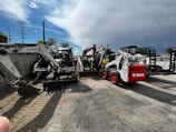 A collection of construction equipment, including multiple Bobcat machines, is parked in an outdoor lot next to a concrete wall. The scene includes excavators and loaders, all lined up on a paved surface under a partly cloudy sky.