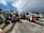 A collection of construction equipment, including multiple Bobcat machines, is parked in an outdoor lot next to a concrete wall. The scene includes excavators and loaders, all lined up on a paved surface under a partly cloudy sky.